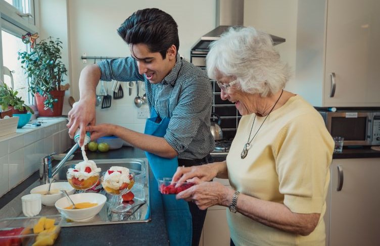 Teenage boy making fruit compote with his grandmother in the kitchen of her home. He is adding squirty cream to the bowls.