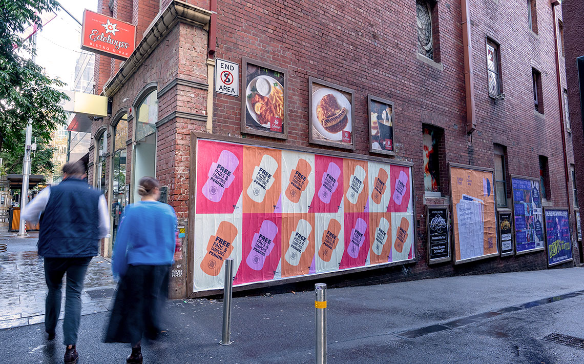 Two people walking down the street with a wall of pads and tampons posters on a building next to them