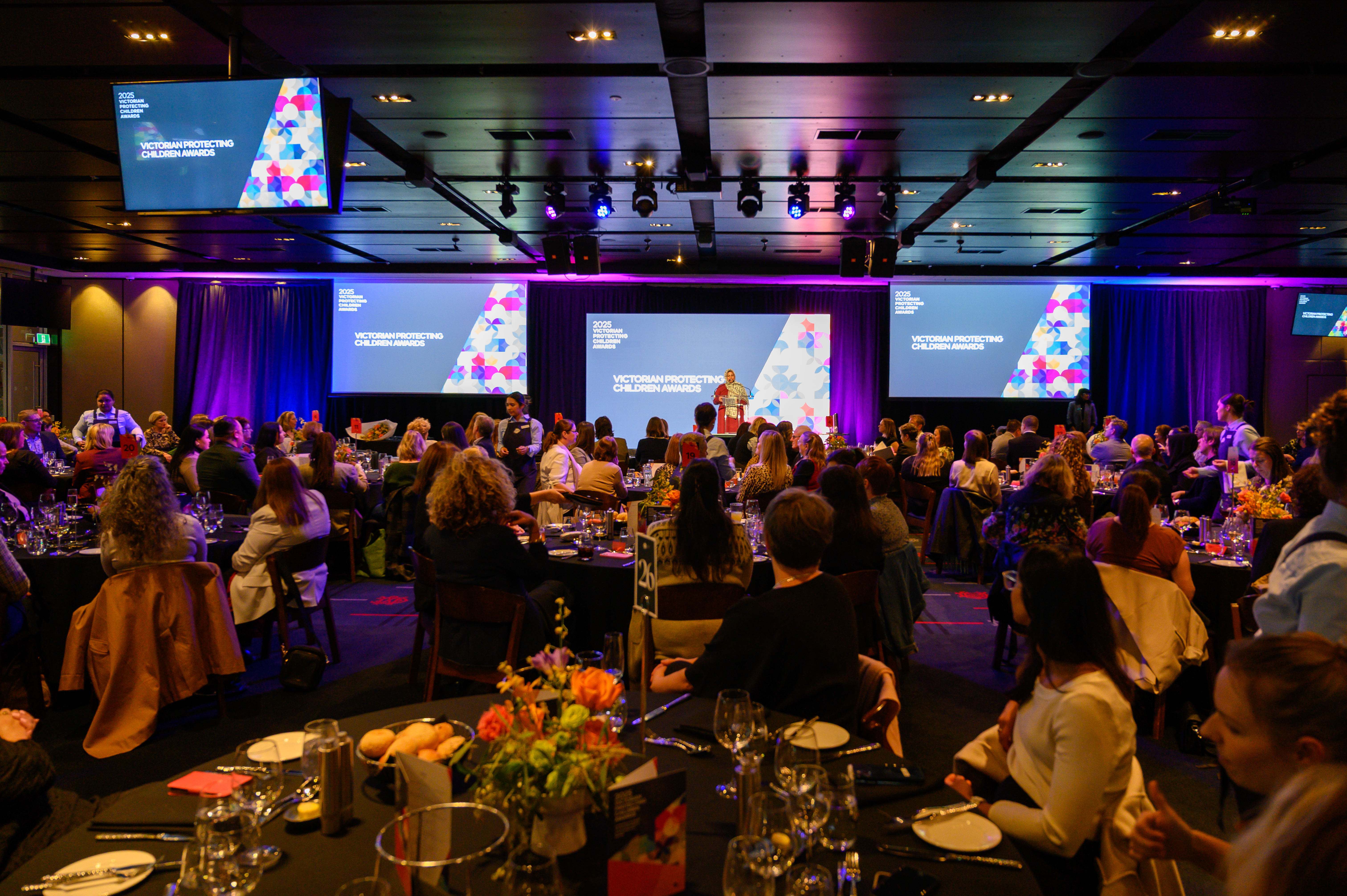 People sitting at tables in the audience of an award ceremony, listening and watching the event.
