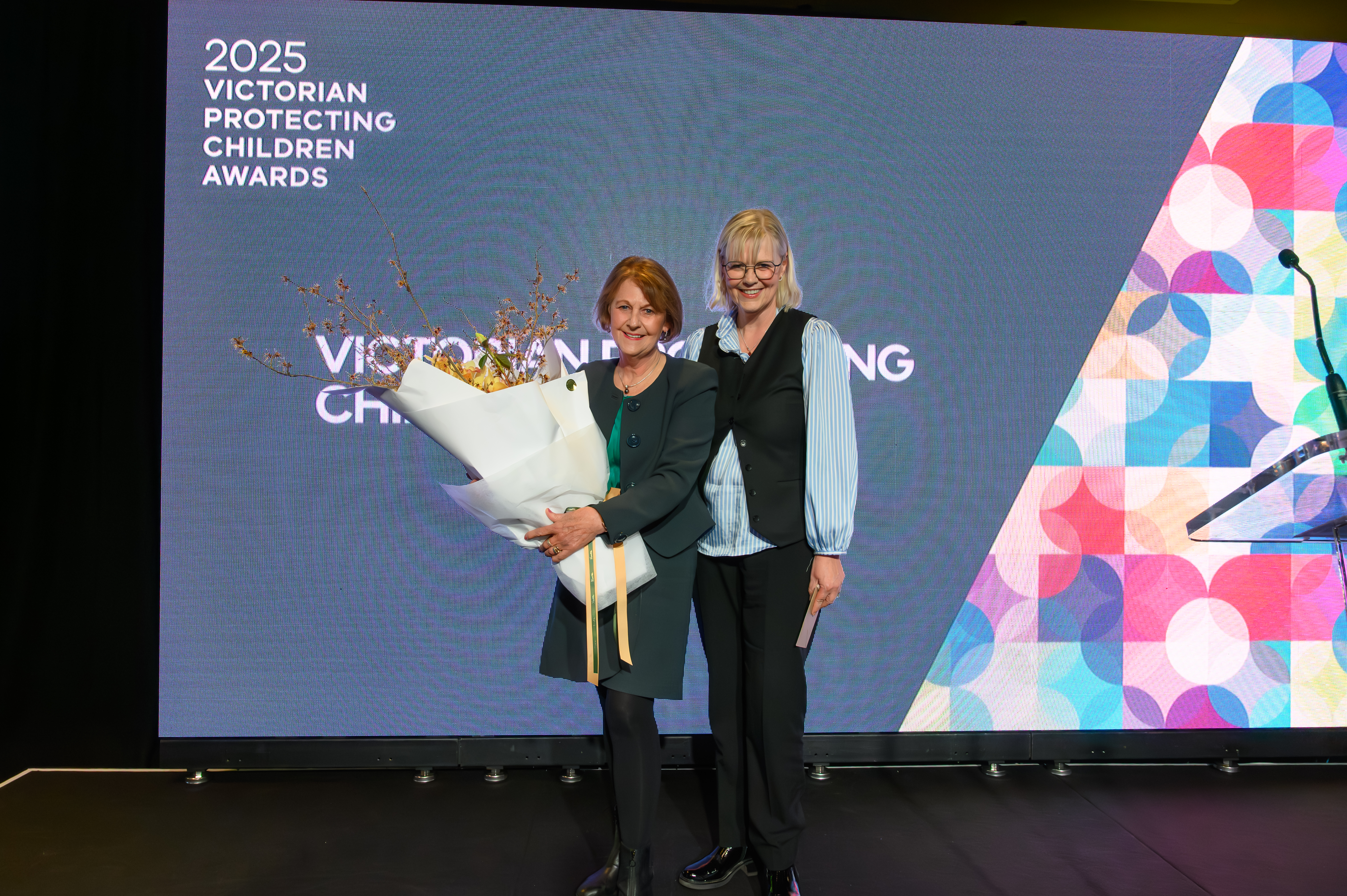Two women standing on stage at an award ceremony; one woman is holding flowers