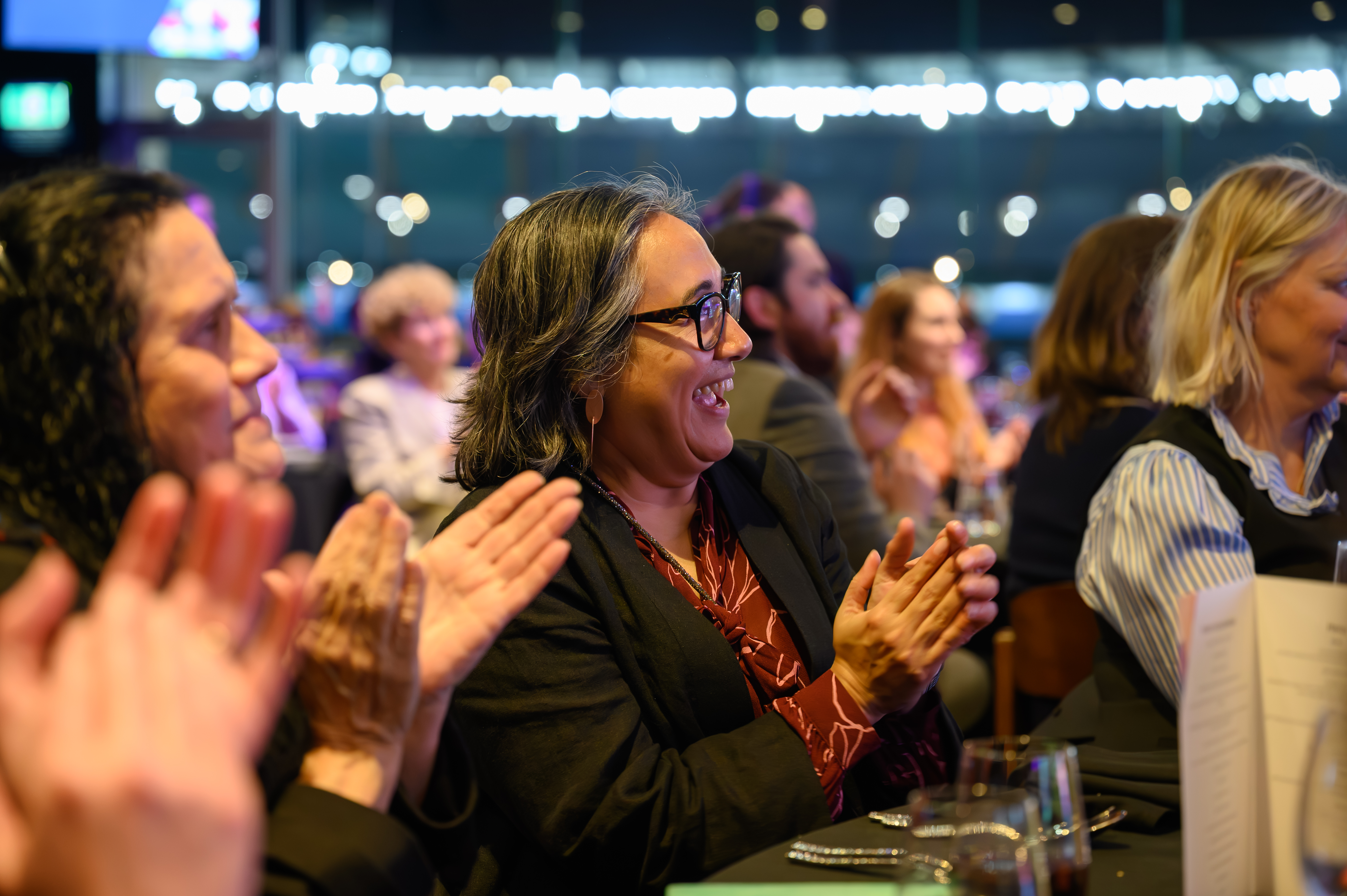 A woman smiling and clapping, sitting in the audience of an awards ceremony