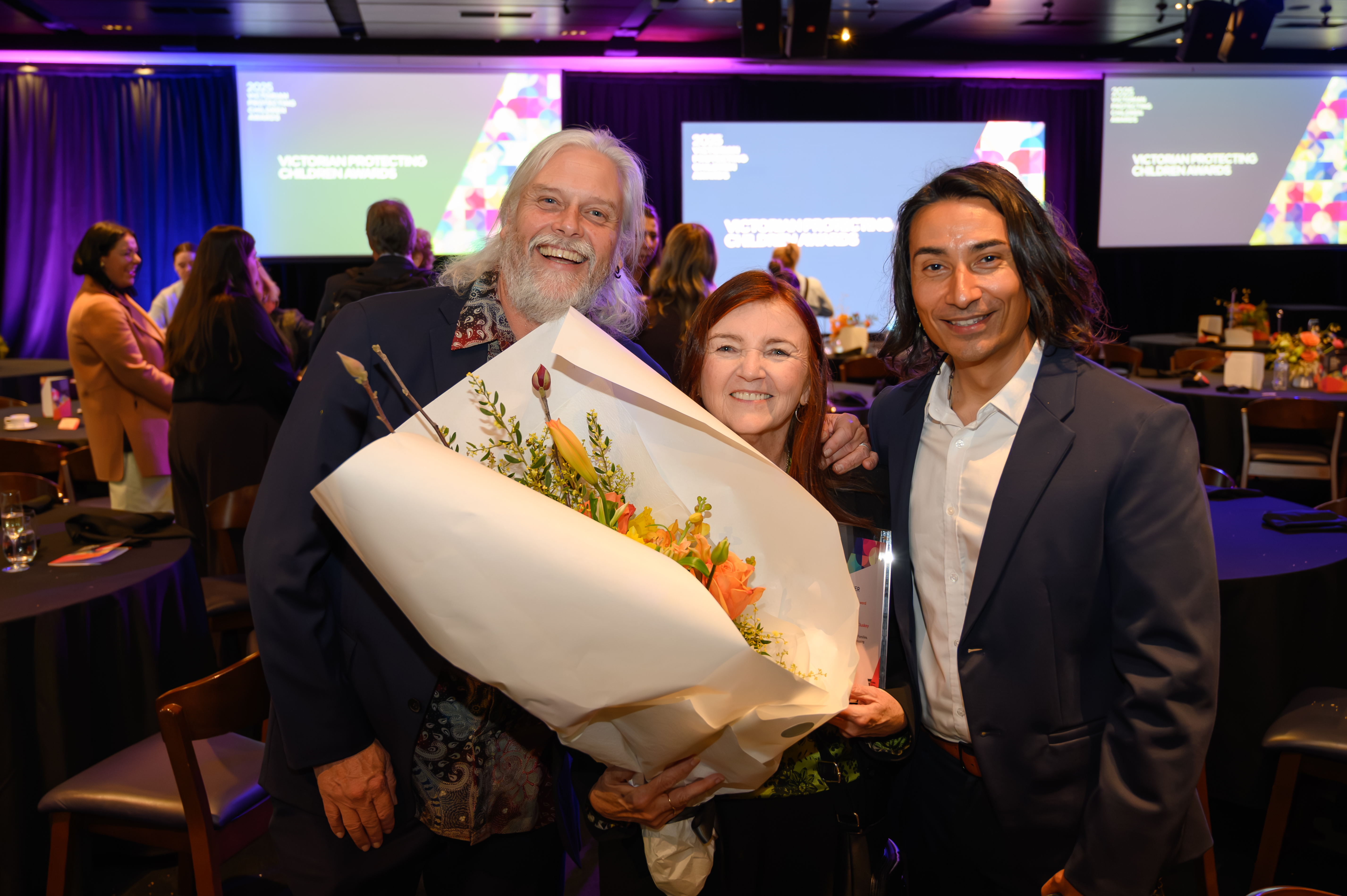 Three people standing together at an award ceremony, the person in the middle is holding flowers.