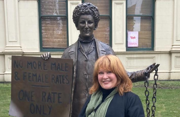 Sculptor Jennifer Mann standing with the statue of Zelda D'Aprano standing in front of the Victorian Trades Hall. She is holding a sign in her right hand that says "No more male and female rates. One rate only". In her left hand is the chain the from around her waist and would be chaining her to the courthouse.